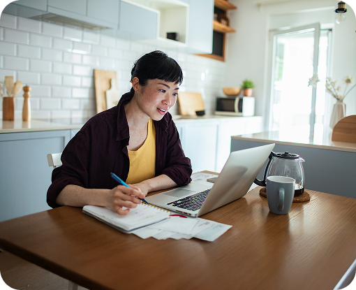 A person writing notes beside a laptop at a dining table